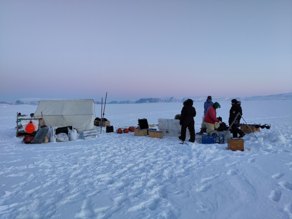 photo showing a small white tent on a snow covered sea ice surface with people dressed in thick warm clothes dropping instruments through a whole in the ice. The sky is a clear blue fading to vioet and pink at sunset